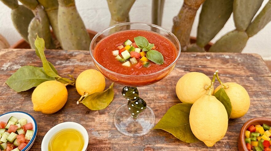 Glasstielschale mit roter Gazpacho, garniert mit gewürfelter Gurke, Paprika und Basilikum; umgeben von frischen Zitronen, Olivenöl und Schälchen mit Gemüsewürfeln.