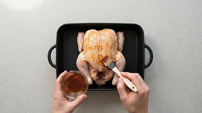 Zwei Hände bestreichen ein rohes Hähnchen in einer schwarzen Auflaufform mit roter Marinade aus einer Glasschale.