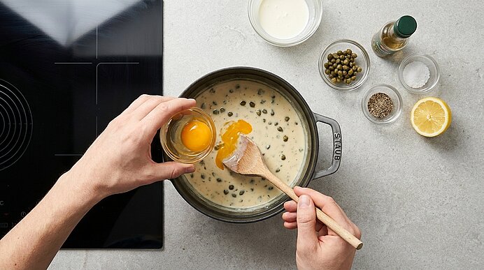 Nahaufnahme von Händen, die ein Ei in einen Topf mit cremiger Kapernsauce geben und die Sauce mit einem Holzlöffel verrühren.