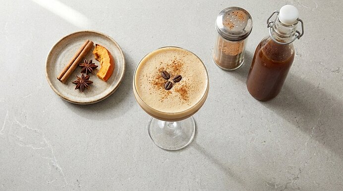 Cocktailglas mit cremigem Kaffee‑Cocktail, garniert mit drei Kaffeebohnen und Zimtpulver; daneben Schale mit Zimtstange, Sternanis und Orangenschale sowie eine Flasche Sirup.