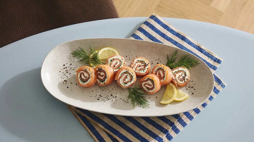 Ovaler Teller mit Lachs-Röllchen gefüllt mit Frischkäse, garniert mit Dill, Zitronenscheiben und schwarzen Pfefferkörnern.