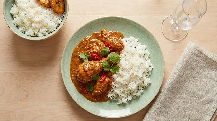 Teller mit Hähnchencurry in reichhaltiger Sauce neben Reis, garniert mit frischen Korianderblättern und dünnen roten Chili‑Scheiben.