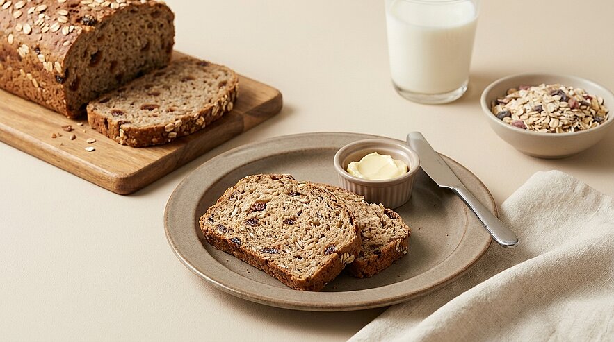 Ein Teller mit zwei Scheiben Brot, das mit Nüssen und Trockenfrüchten gefüllt ist. Neben dem Brot befinden sich ein kleiner Behälter mit Butter und ein Besteck. Im Hintergrund sind ein Glas Milch und eine Schüssel mit Haferflocken sichtbar.