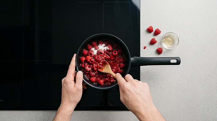 Zwei Hände rühren Himbeeren und Zucker mit einem Holzlöffel in einem schwarzen Stieltopf auf dem Kochfeld um.