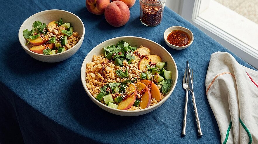 Zwei Schalen mit sommerlichem Getreidesalat aus Bulgur, grob geschnittenen Pfirsichen, Gurkenwürfeln, frischem Koriander und gehackten Nüssen, daneben eine kleine Schale mit Dressing und Besteck.