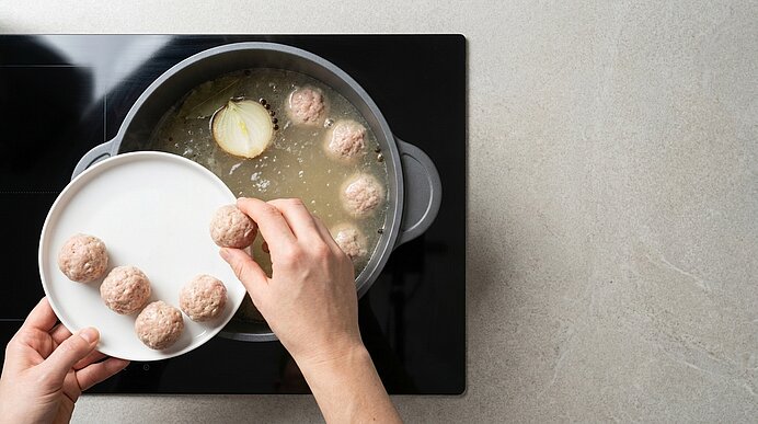 Hände geben rohe Fleischbällchen von einem Teller in einen Topf mit siedender Brühe; eine halbierte Zwiebel schwimmt darin.