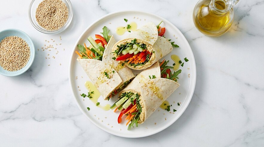 Drei halbierte Tortilla-Wraps auf einem weißen Teller, gefüllt mit frischen Streifen von Karotte, Gurke, roter Paprika und grünen Blättern, mit Sesam bestreut und leichtem Öl.