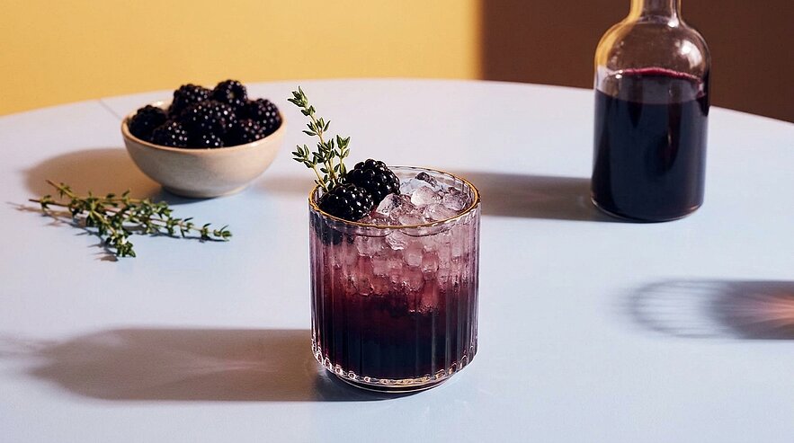 Eisgefülltes Tumbler-Glas mit dunkelviolettem Brombeer-Cocktail, garniert mit frischen Brombeeren und einem Thymianzweig.