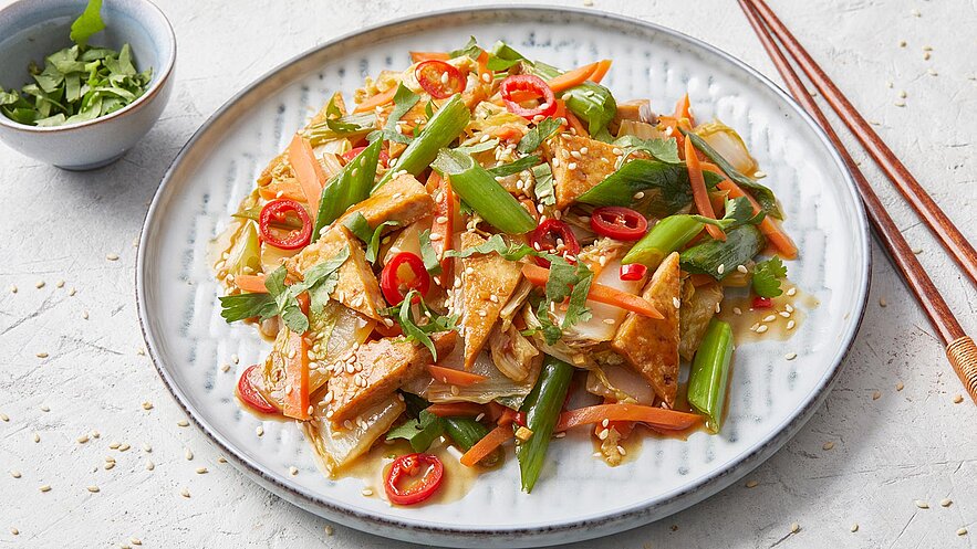 Ein Teller mit buntem Gemüsegericht, bestehend aus gebratenem Tofu, Karotten, Frühlingszwiebeln und Paprika, garniert mit Koriander und Sesam. Es sind auch rote Chili-Scheiben sichtbar. Im Hintergrund befindet sich eine kleine Schüssel mit frischen Kräutern.