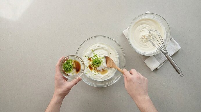 Zwei Hände rühren Magerquark mit Limettenabrieb und Vanille in einer Schüssel. Daneben steht geschlagene Sahne.
