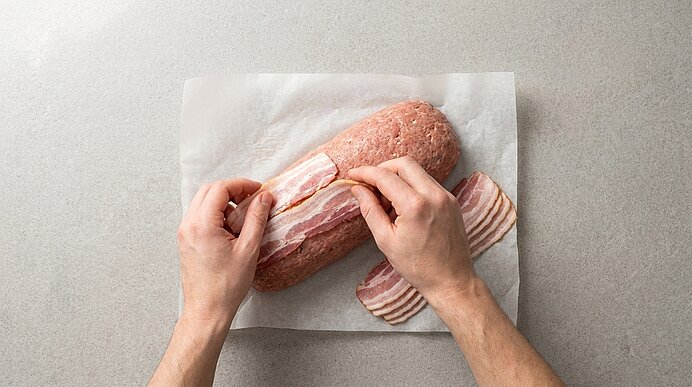 Zwei Hände legen Speckstreifen auf eine längliche rohe Hackfleischrolle auf Backpapier, um einen Speckmantel zu formen.