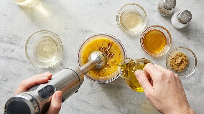 Zwei Hände bereiten ein Senf‑Vinaigrette zu: Stabmixer in einer Schüssel mit grobem Senf, während Olivenöl eingegossen wird; daneben kleine Schälchen mit Essig, Honig und Gewürzen.