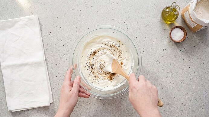 Zwei Hände verrühren Schmand, Crème fraîche und Gewürze mit einem Holzlöffel in einer Glasschüssel.