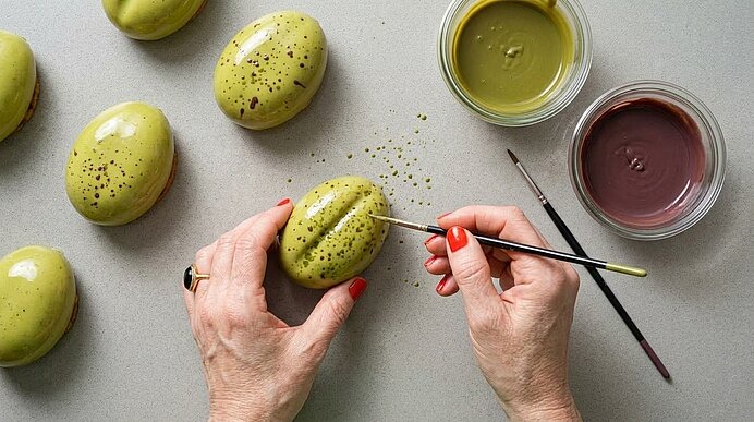 Zwei Hände bemalen ein pistazienförmiges Dessert mit Pinsel, daneben Schälchen mit grüner und brauner Kuvertüre.