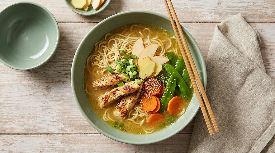 Eine große Schüssel mit Nudelsuppe, garniert mit Hähnchenstreifen, frischen Kräutern, Karotten, Zuckerschoten und Ingwerscheiben. Neben der Schüssel befindet sich eine kleine Schale mit Ingwer und ein Essstäbchen auf der Suppe. Der Hintergrund ist hell und aus Holz.