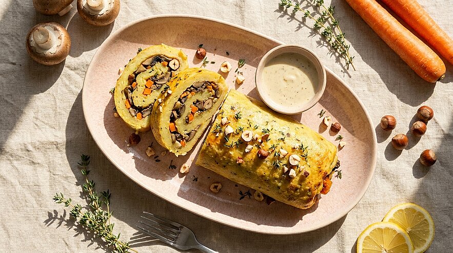 Gebackene Gemüse‑Roulade auf ovalem Teller, in Scheiben geschnitten, serviert mit einer kleinen Schale Sauce und garniert mit gehackten Haselnüssen und frischen Kräutern.