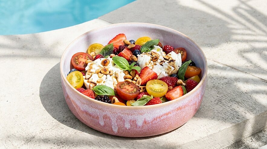 Rosafarbene Schale mit sommerlichem Salat: zwei Burrata-Kugeln, gelbe und rote Kirschtomaten, Erdbeeren und Beeren, frische Basilikumblätter, gehobelte Nüsse und Olivenöl.