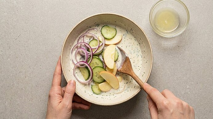 Hände rühren in einer Schüssel Gurkenscheiben, Apfelscheiben und rote Zwiebelringe in cremigem Dressing mit einem Holzlöffel; daneben kleine Schale mit Dressing.