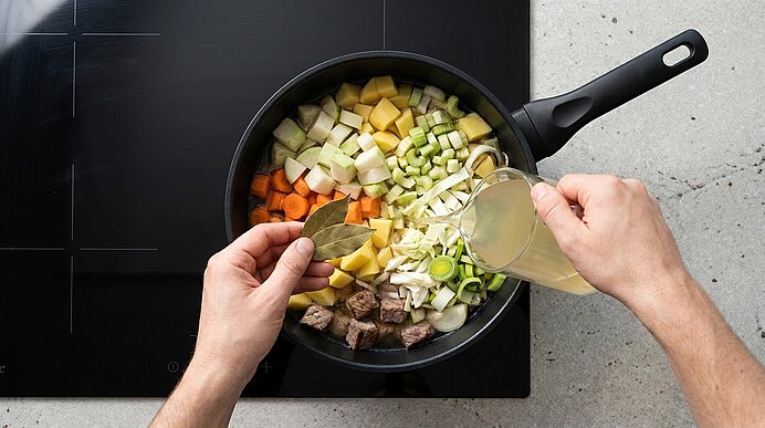 Zwei Hände geben ein Lorbeerblatt und gießen Brühe in eine Pfanne mit gewürfeltem Gemüse (Karotten, Kartoffeln, Lauch, Zwiebeln) und Fleischwürfeln auf dem Herd.