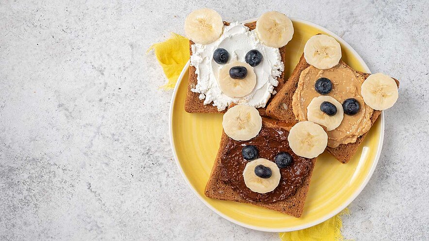 Drei mit Lebensmitteln dekorierte Brotscheiben in Form von Bärenköpfen auf einem gelben Teller. Die Brote sind mit verschiedenen Aufstrichen (frischkäse, Erdnussbutter, Schokoladencreme) belegt und mit Bananen- und Heidelbeerstücken verziert, die als Augen und Ohren dienen.