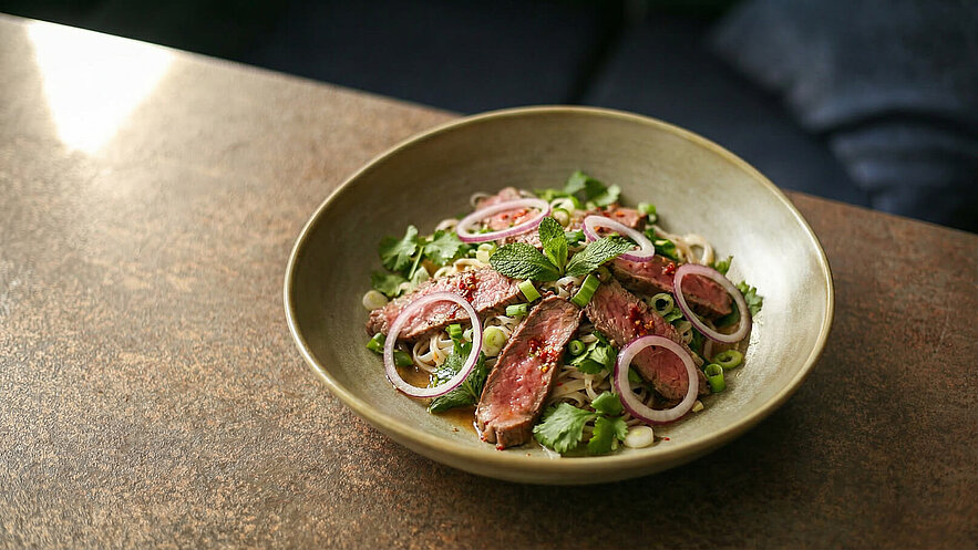 Eine Schüssel mit Nudelsalat, garniert mit zarten Rindersteaks, roten Zwiebelringen, frischem Koriander und Minze. Grüne Zwiebelstücke und scharfe Gewürze sind ebenfalls sichtbar. Der Salat ist auf einer Holzoberfläche platziert.