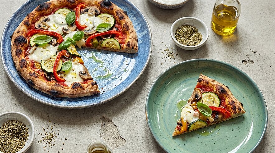 Eine frisch gebackene Pizza mit roten Paprika, Zucchini, Pilzen und Mozzarella, garniert mit frischem Basilikum. Ein Stück der Pizza ist auf einem grünen Teller angerichtet. Um die Pizza herum sind Gewürze und Olivenöl in kleinen Schalen dekoriert.