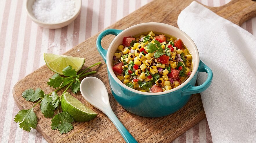 Türkisfarbene Schüssel mit Mais-Salsa: gerösteter Mais, gewürfelte Tomaten und Paprika, frischer Koriander und Kräuter, dazu Limettenviertel und Löffel auf einem Holzbrett.