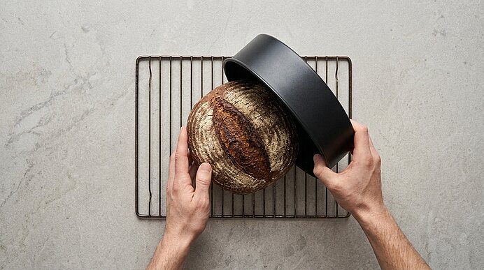 Zwei Hände heben eine runde schwarze Backform von dem gebackenen Roggensauerteigbrot auf einem Abkühlgitter ab.