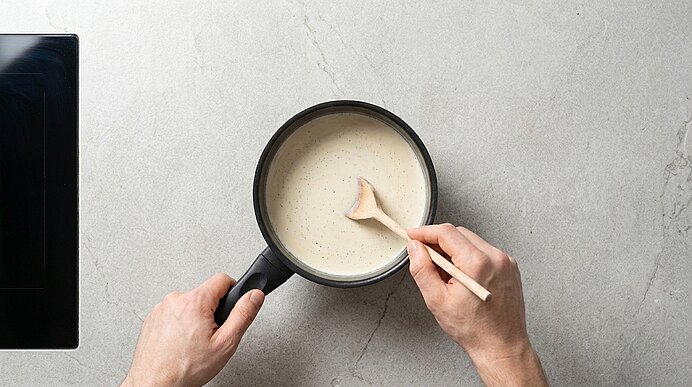 Zwei Hände halten einen kleinen Topf und rühren mit einem Holzlöffel eine cremige helle Sauce auf einer Arbeitsfläche.