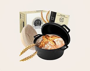 Ein gusseiserner Topf mit frisch gebackenem Brot, das eine goldene Kruste hat, steht im Vordergrund. Im Hintergrund sind eine Schachtel mit Rezepten sowie einige Weizenkörner zu sehen. Der Fokus liegt auf dem Brot und dem Topf.