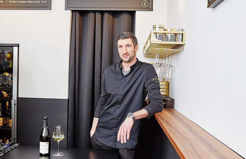 Ein Mann steht an einer hölzernen Theke in einem Lokal. Er trägt ein schwarzes Hemd und lächelt. Vor ihm steht ein Glas mit Getränk und eine Flasche. Im Hintergrund sind eine Getränkeflaschen und ein schwarzes Vorhang zu sehen.