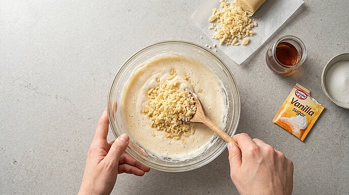 Zwei Hände rühren mit einem Holzlöffel Teig in einer Glasschüssel, dabei werden grob gehackte Stücke (z. B. Nüsse oder weiße Schokolade) in den Teig gegeben.