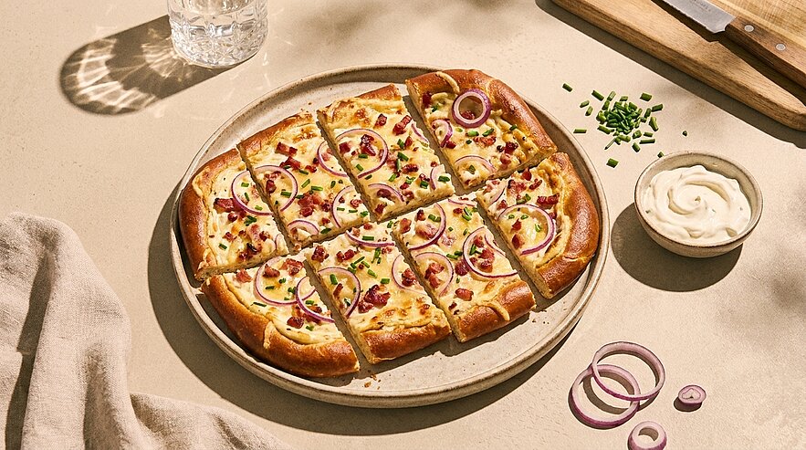 Auf einem Teller liegt eine in neun Quadrate geschnittene Pizza mit heller Sauce, roten Zwiebelringen, Speckstückchen und gehacktem Schnittlauch; daneben eine kleine Schale mit cremigem Dip.