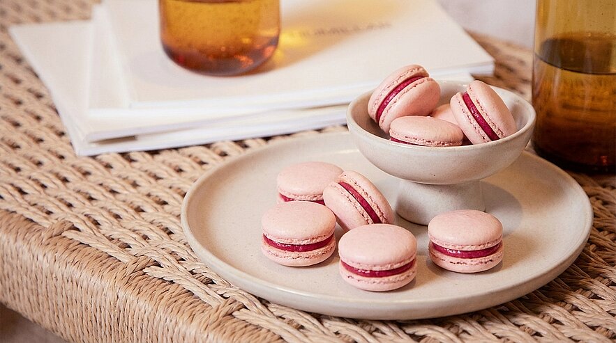 Ein Teller mit rosa Macarons steht auf einem geflochtenen Tisch. Einige Macarons liegen außerhalb der Schüssel, während andere in einer kleinen Schüssel präsentiert werden. Im Hintergrund sind einige Stapel weißer Blätter und ein Glas sichtbar.