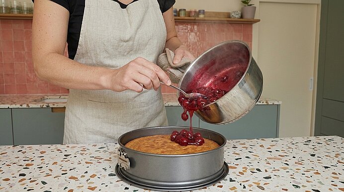 Person in Schürze gießt frisch gekochte Kirschen aus einem Topf als Fruchtbelag auf einen gebackenen Kuchen in einer Springform.