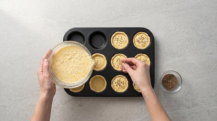 Zwei Hände giessen Käsemasse aus einer Glasschüssel in Teigförmchen im Muffinblech und bestreuen sie mit Kümmel.