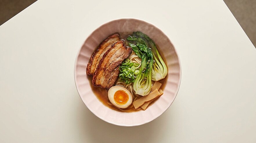 Eine rosa Schüssel mit Ramen-Nudeln, garniert mit gegrilltem Fleisch, einem halben gekochten Ei, Pak Choi und Frühlingszwiebeln. Das Gericht ist appetitlich angerichtet und zeigt eine Mischung aus verschiedenen Zutaten.
