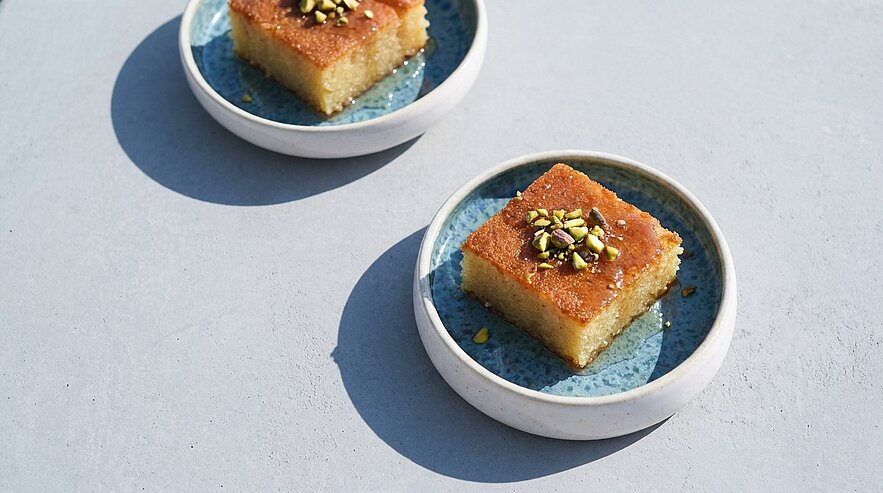 Zwei kleine Teller mit einem Stück saftigen Baklava. Das Gebäck ist goldbraun und mit gehackten Pistazien bestreut. Die Teller haben einen blauen Rand und stehen auf einer hellen Oberfläche.