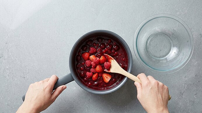 Top‑Ansicht: Hände rühren mit einem Holzlöffel in einem Topf ein rotes Beerenkompott aus Erdbeeren und Himbeeren.