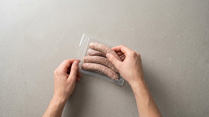 Zwei Hände halten eine Vakuumverpackung mit vier rohen Bratwürsten auf einem hellgrauen Untergrund.
