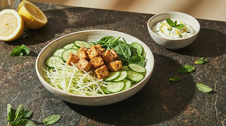 Schale mit frischem Salat: gewürfelter, gebratener Tofu, Gurkenscheiben, feine Kohlstreifen und Blattspinat, dazu ein Schälchen Joghurt-Dip und Zitronenhälften.