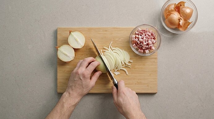 Hände schneiden eine Zwiebel in feine Streifen auf einem Holzbrett; daneben eine Schüssel mit gewürfeltem Speck und eine Schüssel mit Zwiebelhäuten.