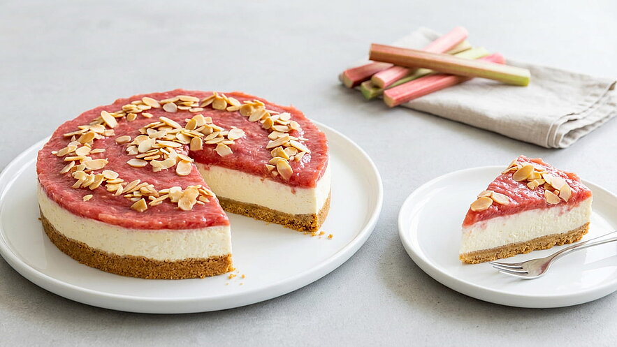 Ein Käsekuchen mit einer schneeweißen Füllung und einer roten Rhabarber-Toppings, garniert mit gerösteten Mandeln. Ein Stück des Kuchens liegt neben dem ganzen Kuchen auf einem Teller, während Besteck daneben platziert ist.