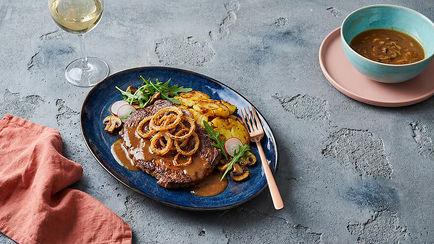 Ein tiefes Teller mit einem Stück gebratenem Fleisch, übergossen mit Soße und garnieren mit Rucola, Radieschen und Pilzen. Neben dem Fleisch liegen goldene Bratkartoffeln. Im Hintergrund steht ein Glas Weißwein und eine Schüssel mit einer Brühe.