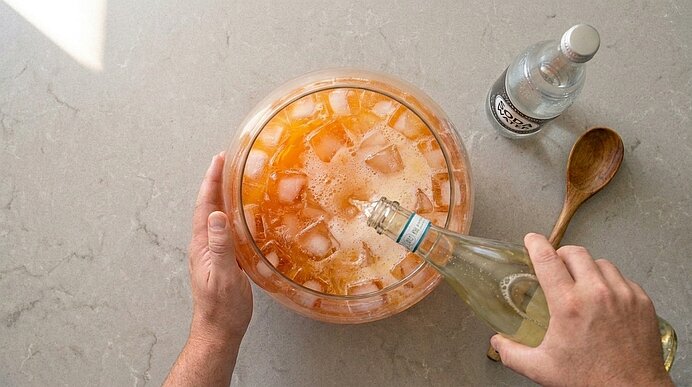 Zwei Hände gießen Prosecco in eine große Glasschüssel mit eisgekühlter oranger Bowle auf einer hellgrauen Arbeitsplatte.