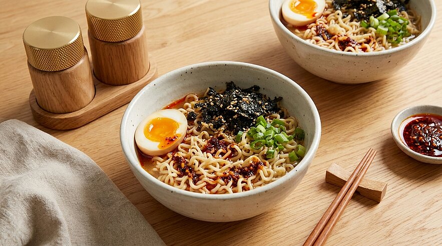 Zwei Schalen Ramen-Nudeln mit halbierten weich gekochten Eiern, gehackten Frühlingszwiebeln, zerrissenem Nori, Sesam und Chili-Öl; Essstäbchen liegen daneben.