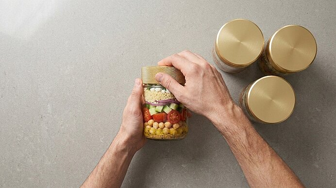 Zwei Hände verschließen ein Glas mit geschichtetem Salat (Mais, Kirschtomaten, Gurke, rote Zwiebel, Blattsalat, Käse), daneben drei weitere Gläser mit goldenen Deckeln auf grauer Oberfläche.