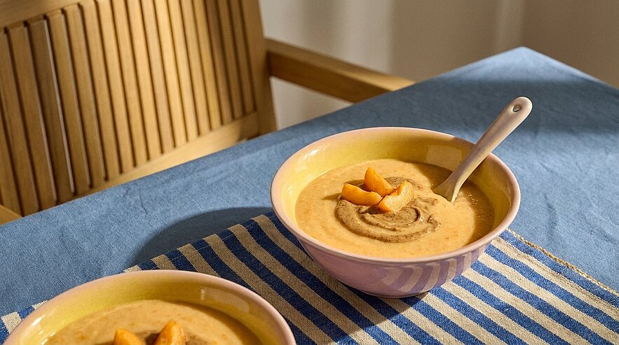Zwei Schalen mit cremiger orangefarbener Suppe, eine mit einem Löffel, beide mit Fruchtstücken garniert und auf einem gestreiften Tischtuch arrangiert.