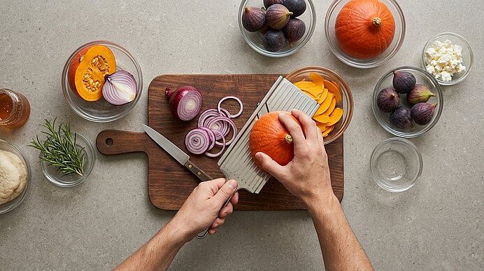 Zwei Hände hobeln Hokkaido-Kürbis mit einem Gemüsehobel auf einem Holzbrett, auf dem Zwiebelringe liegen.