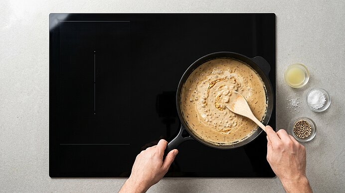 Hände rühren mit Holzlöffel eine cremige Sauce in einem schwarzen Topf auf einem Induktionskochfeld; rechts kleine Schälchen mit Gewürzen.